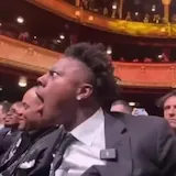 Man in suit reacts with shock in theater audience at 0:03