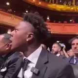 Man in suit reacts with shock in theater audience at 0:06