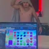 A shirtless man in a towel hood throws his arms open behind a glass at 0:11