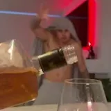 A shirtless man in a towel hood throws his arms open behind a glass at 0:03