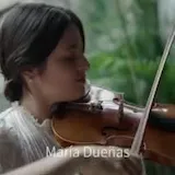 Maria Dueñas plays violin alone in a garden, then with a full orchestra at 0:01