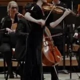 Maria Dueñas plays violin alone in a garden, then with a full orchestra at 0:35