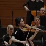 Maria Dueñas plays violin alone in a garden, then with a full orchestra at 0:23