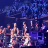 A packed crowd stands in blue light and films the show at an awards at 0:01