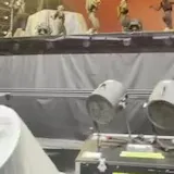 Dancers move on a lit stage as a camera peers past backstage equipment at 0:02