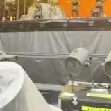 Dancers move on a lit stage as a camera peers past backstage equipment at 0:01