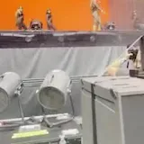 Dancers move on a lit stage as a camera peers past backstage equipment at 0:05